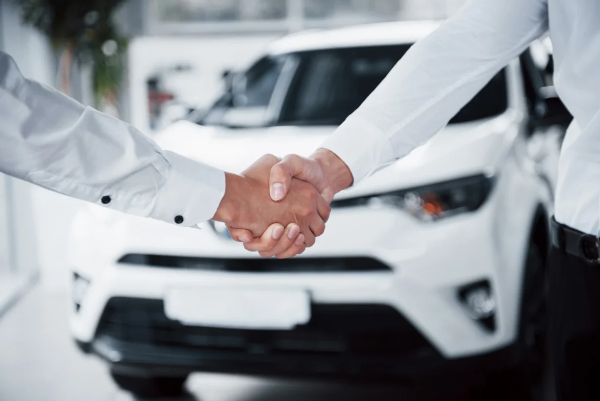 Two people shaking hands in front of a white car at a dealership, 247lemonlaw