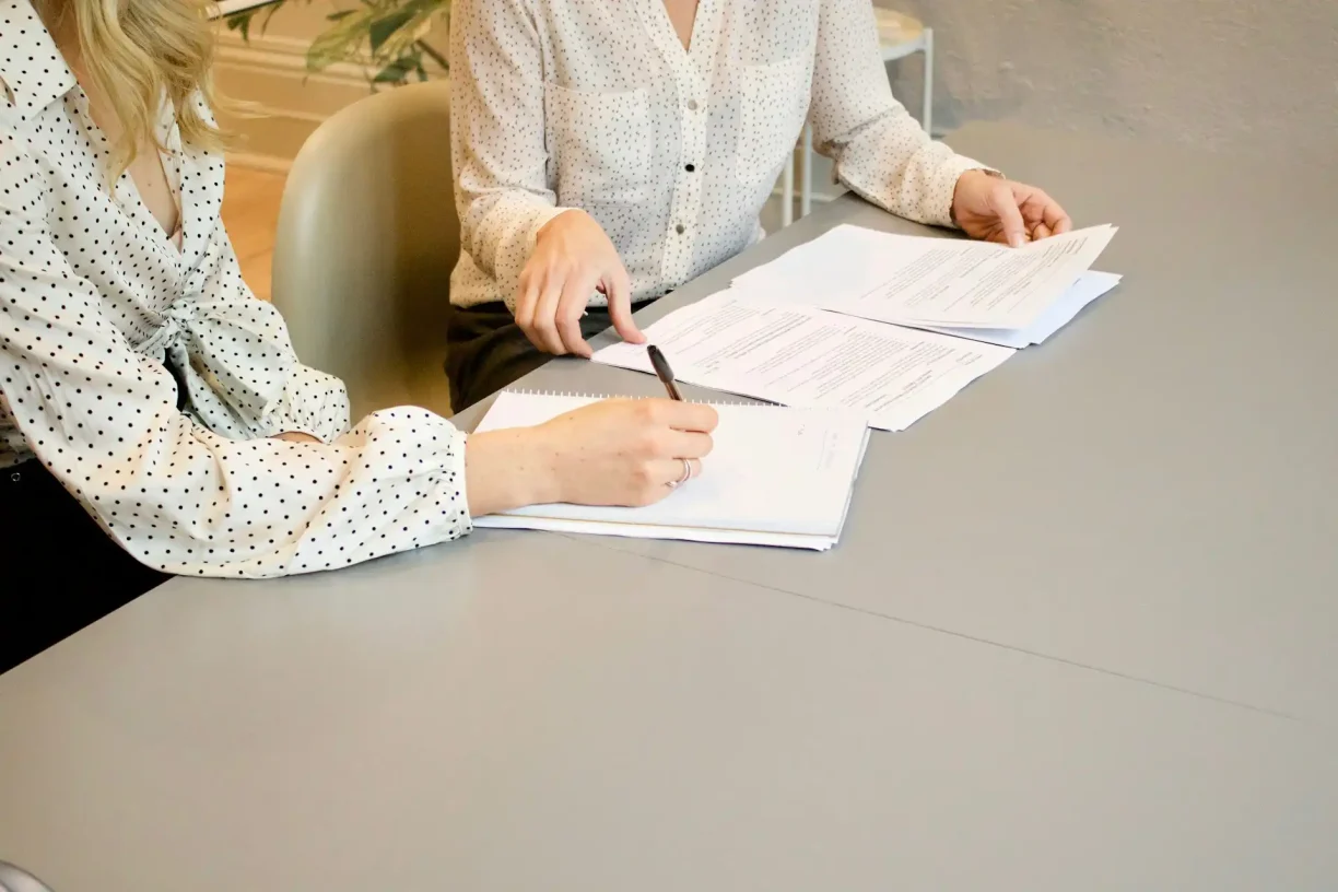 Two women collaborating, one writing and another reviewing documents at a table, 247lemonlaw