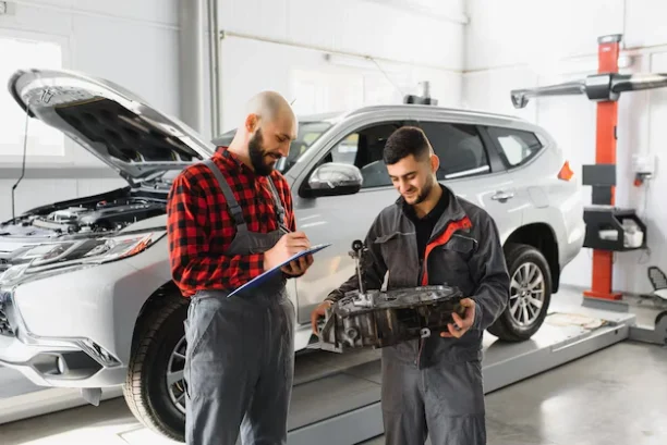 Two mechanics reviewing a faulty car part and documenting repairs beside a vehicle in the workshop.
